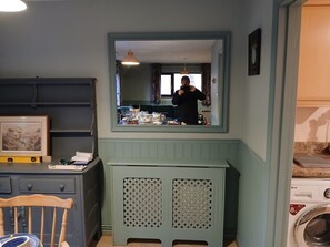 A compact kitchen with a built-in washing machine, a mirror above the sink, and a wooden chair. - Croydon Cottage (Minehead)
