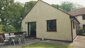 Una casa de un piso con techo inclinado, un patio con mesa y sillas, y un pequeño jardín.