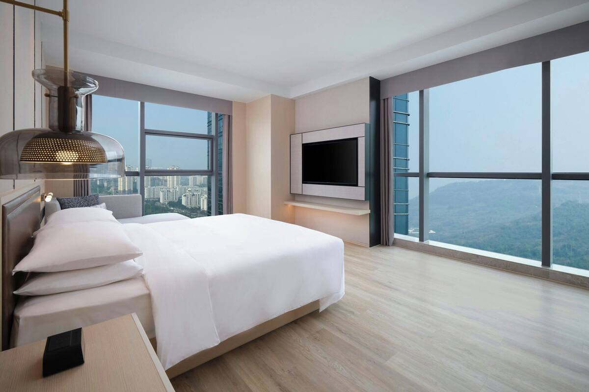 A modern hotel room with a large bed, a flat-screen TV, and a city view through large windows.