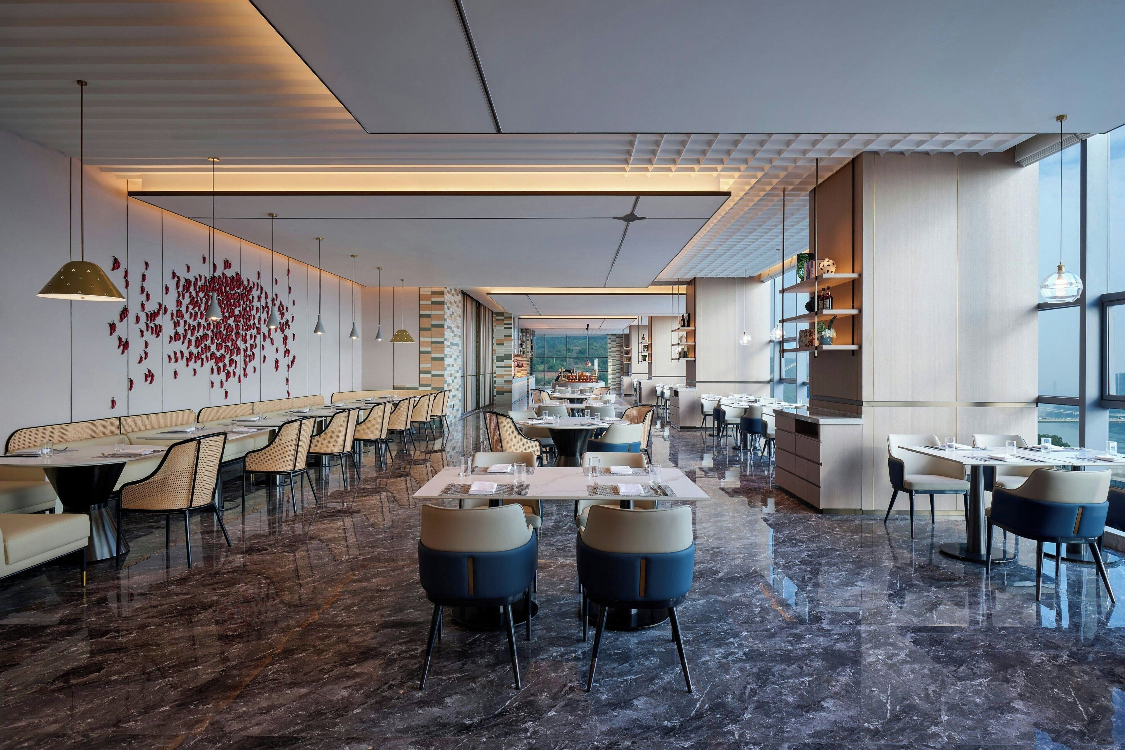 Un restaurant spacieux avec de hauts plafonds, un sol en marbre et des luminaires modernes. Les tables sont disposées pour accueillir les convives, et on peut apercevoir des coins salons avec des fauteuils confortables.