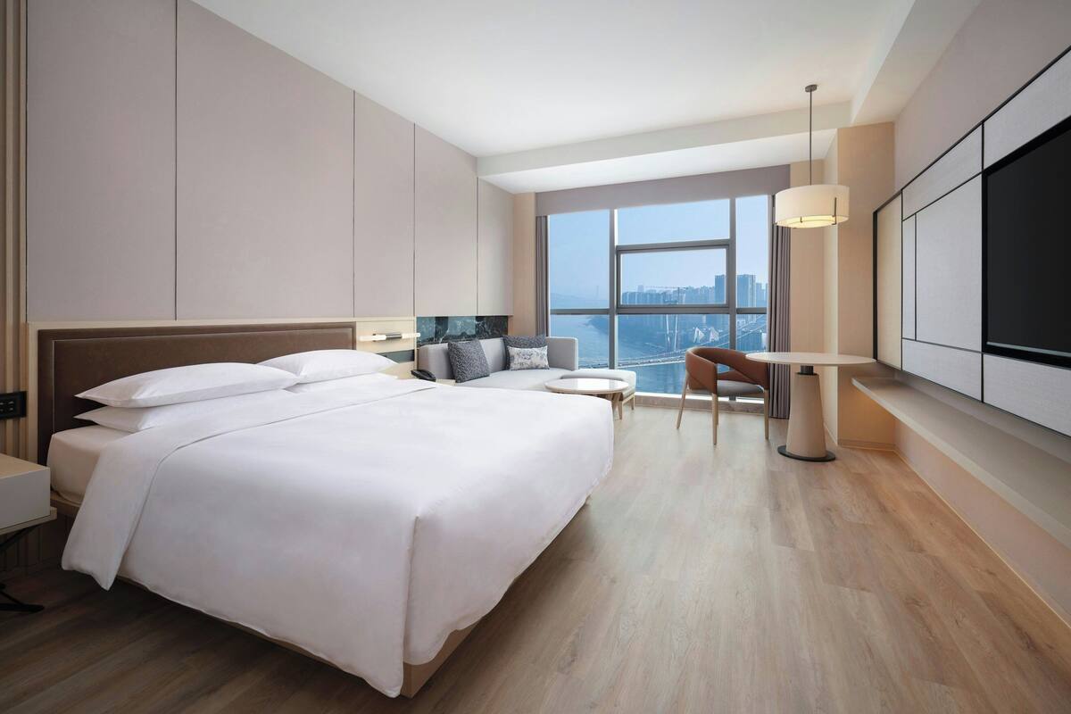 A modern hotel room with a large bed, a desk, a chair, and a large window offering a city view.