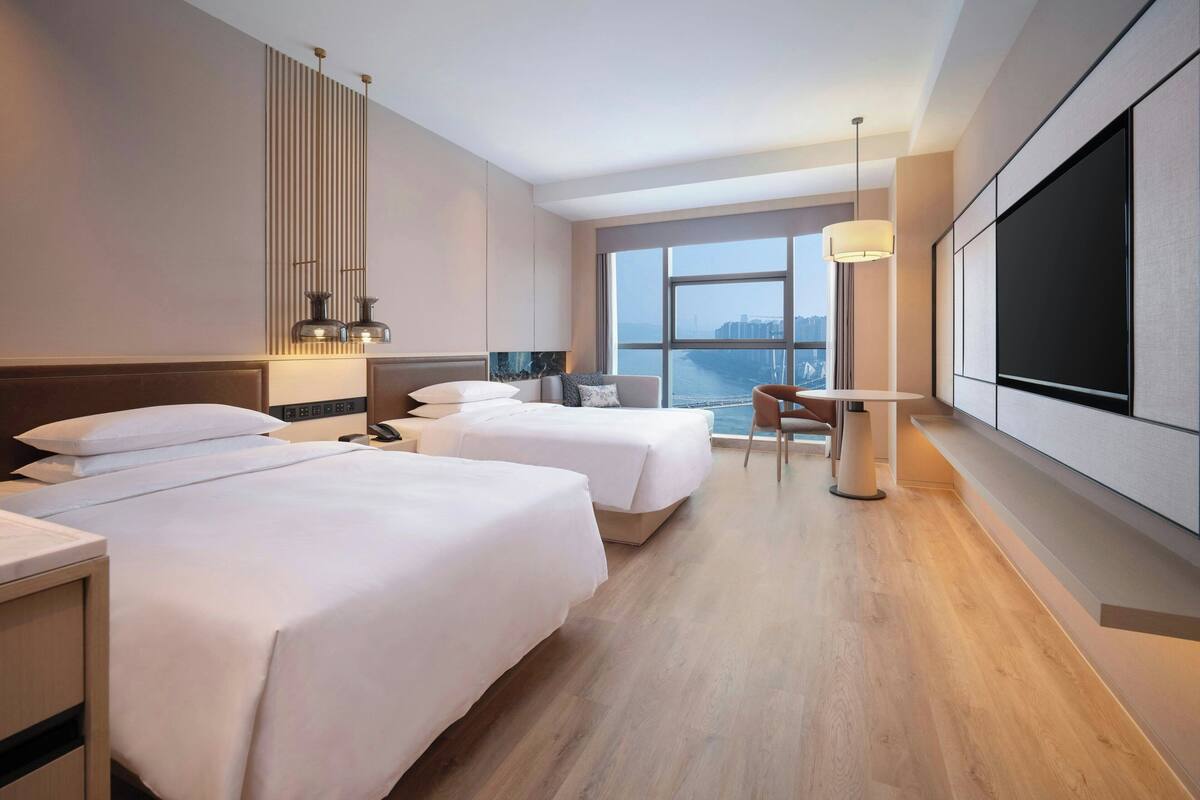 A hotel room with two beds, a desk, a chair, a TV, and a large window with a city view.
