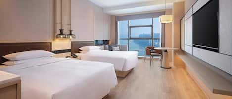 Ein Hotelzimmer mit zwei Betten, einem Schreibtisch, einem Stuhl, einem Fernseher und einem großen Fenster mit Blick auf die Stadt.