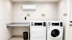 A laundry room with two front-loading washing machines, a small trash bin, a utility wall with a wall-mounted air conditioner, and a linen storage sign. - Springhill Suites By Marriott Madison Fitchburg (Madison)