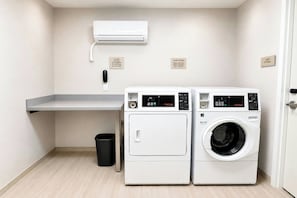 A laundry room with two front-loading washing machines, a small trash bin, a utility wall with a wall-mounted air conditioner, and a linen storage sign. - Springhill Suites By Marriott Madison Fitchburg (Madison)