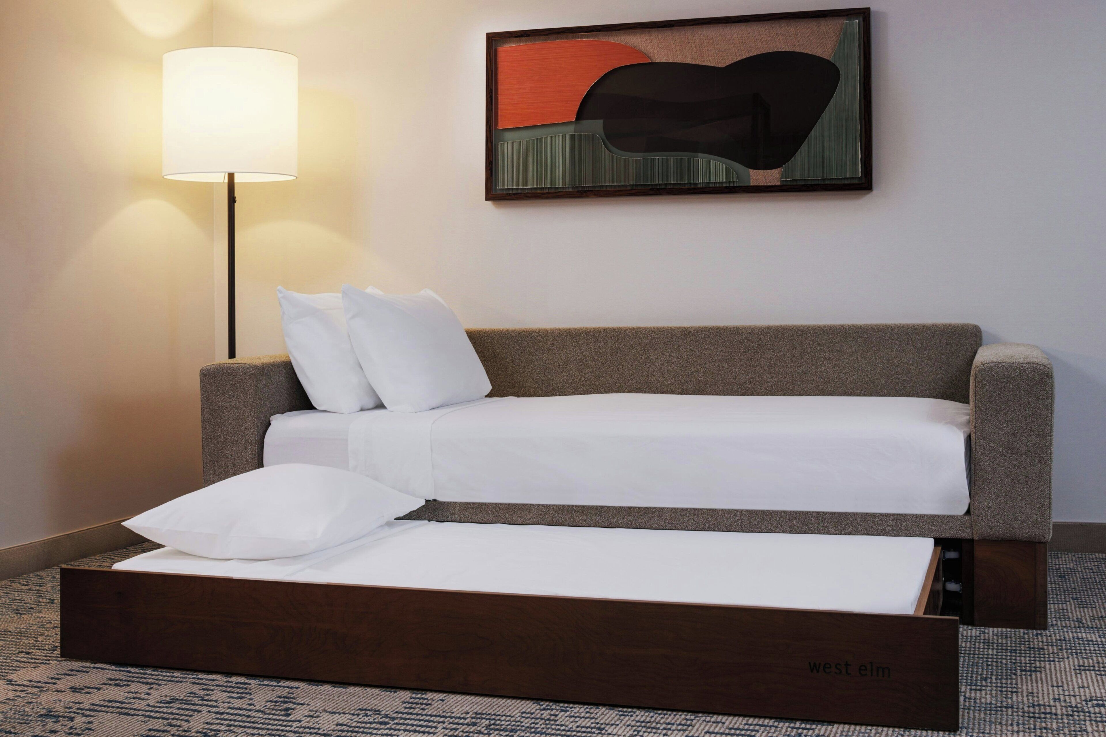A hotel room with a sofa bed, a standing lamp, and a painting on the wall.