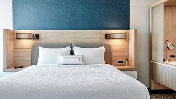 A hotel room with a large bed, two bedside tables, a nightstand, a wall-mounted lamp, and a wooden wardrobe. - Springhill Suites By Marriott Madison Fitchburg (Madison)