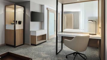 A hotel room with a wooden wardrobe, a flat-screen TV, a white bedside table, a chair, and a desk with a lamp.