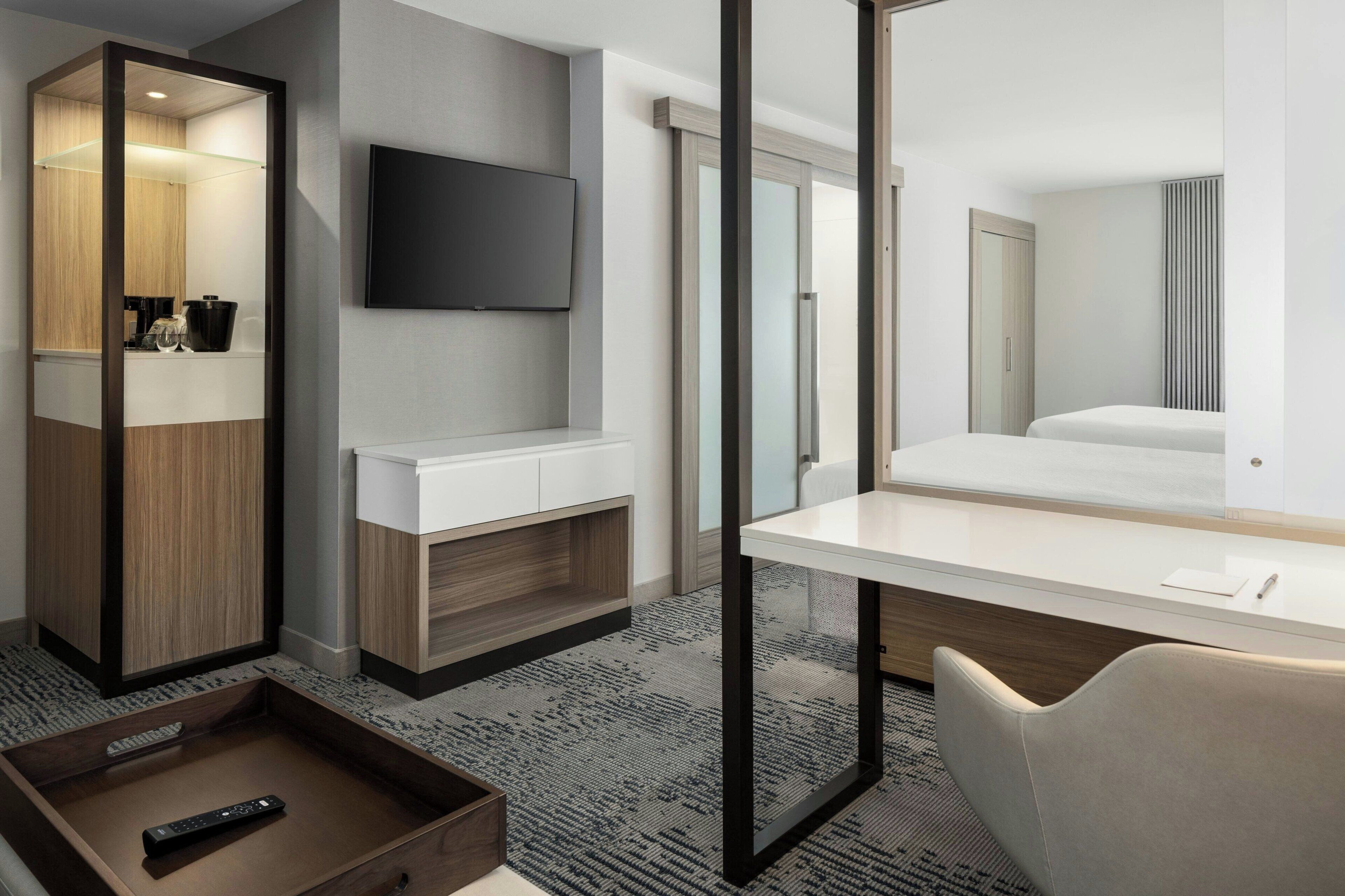 A hotel room with a large mirror, a flat-screen TV mounted on the wall, a bed with a white headboard, a desk with a chair, and a small wooden side table with a remote control.