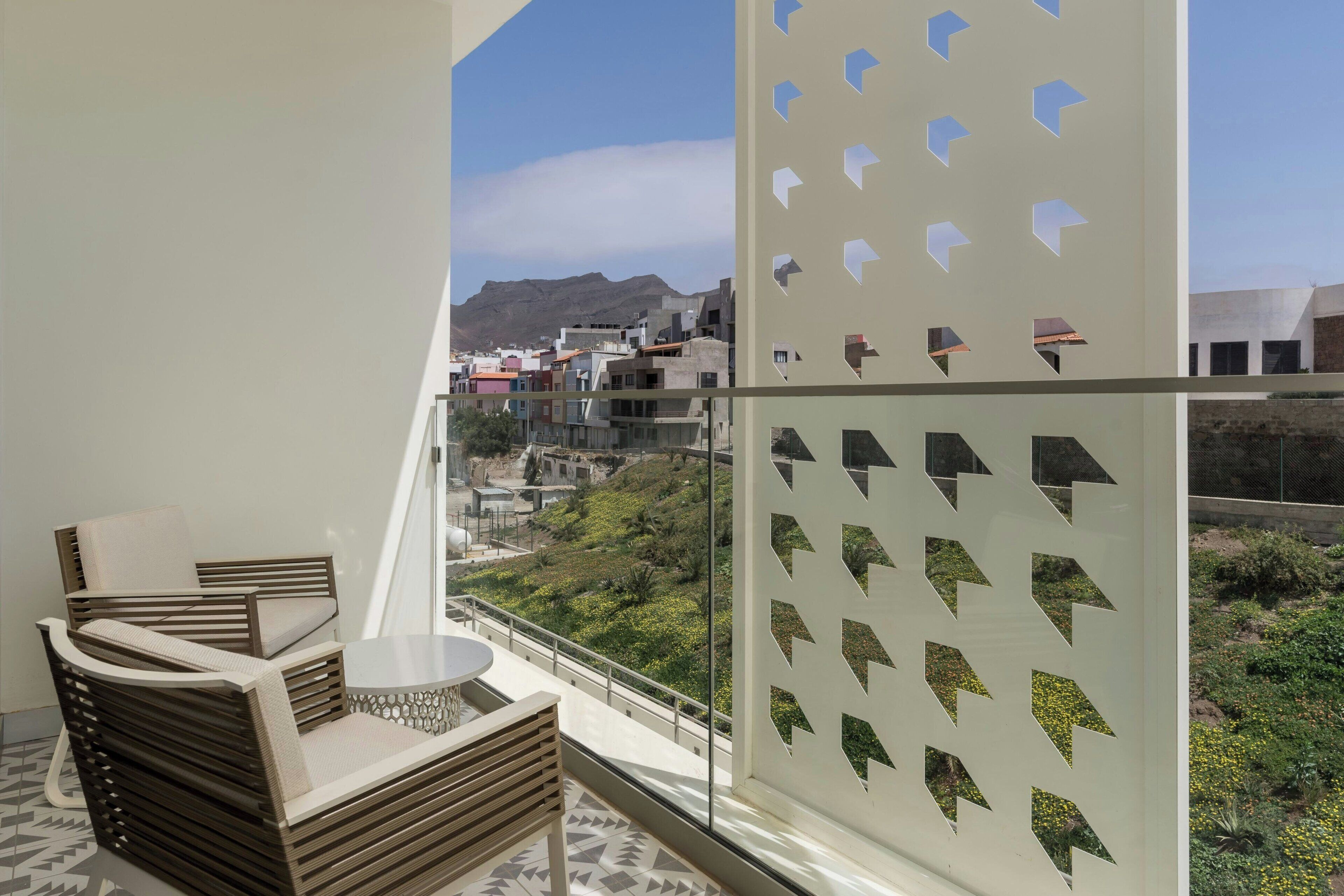 Un balcón con vistas a un pueblo en una colina, una silla de madera, una mesita y un panel decorativo blanco.