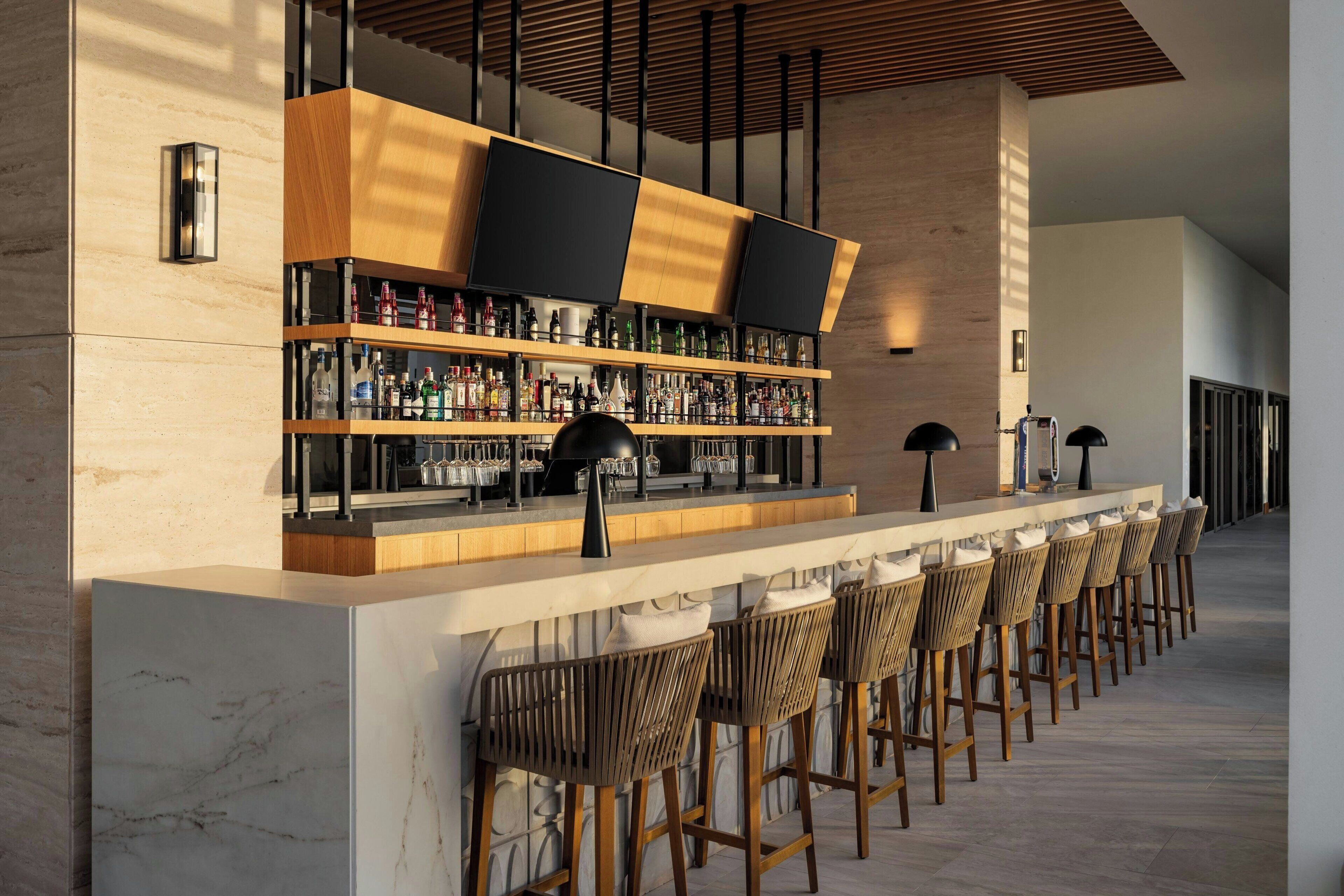 Una zona de bar con una barra larga, varias estanterías llenas de botellas y distintos taburetes.