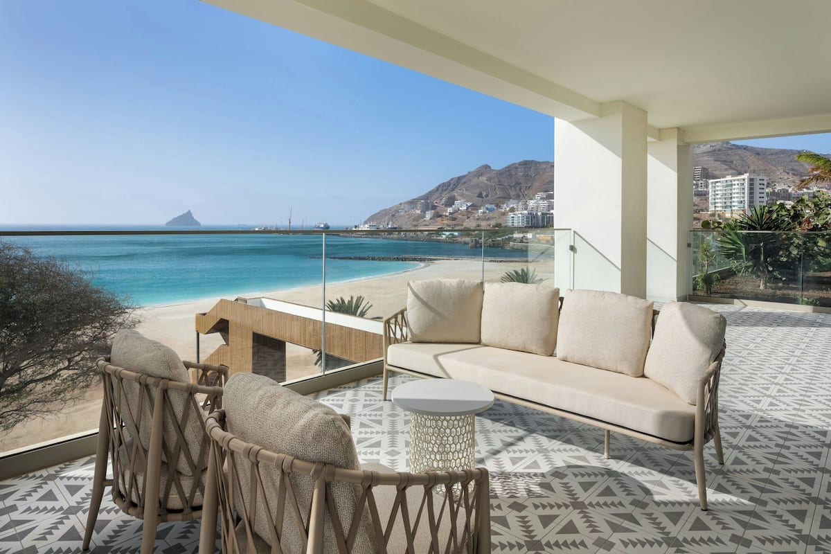 Un balcon agrémenté d’un canapé, de chaises et d’une petite table, offrant une vue sur une plage et des montagnes.