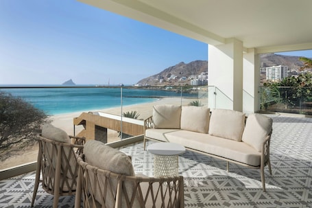 A balcony with a sofa, chairs, and a small table, overlooking a beach and mountains.