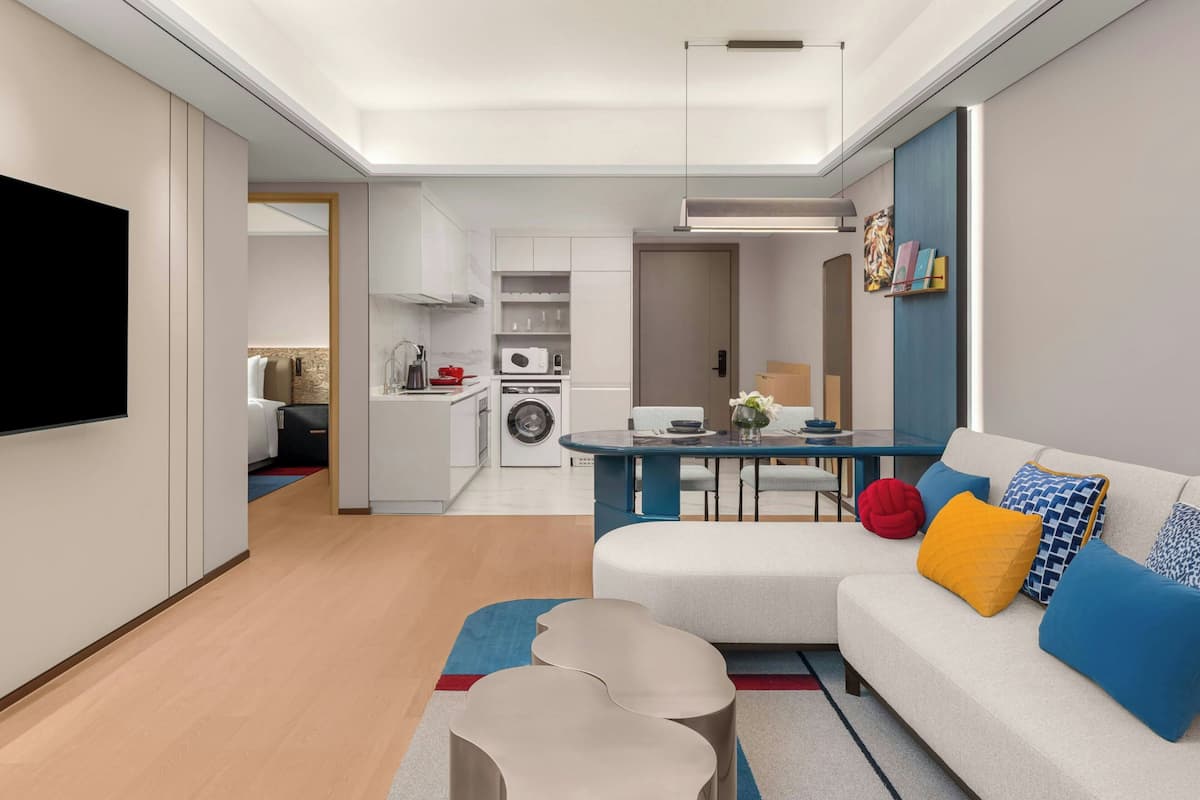 Una sala de estar moderna con un sofá, una mesa de centro y un televisor. Hay una zona de cocina con armarios y un microondas.