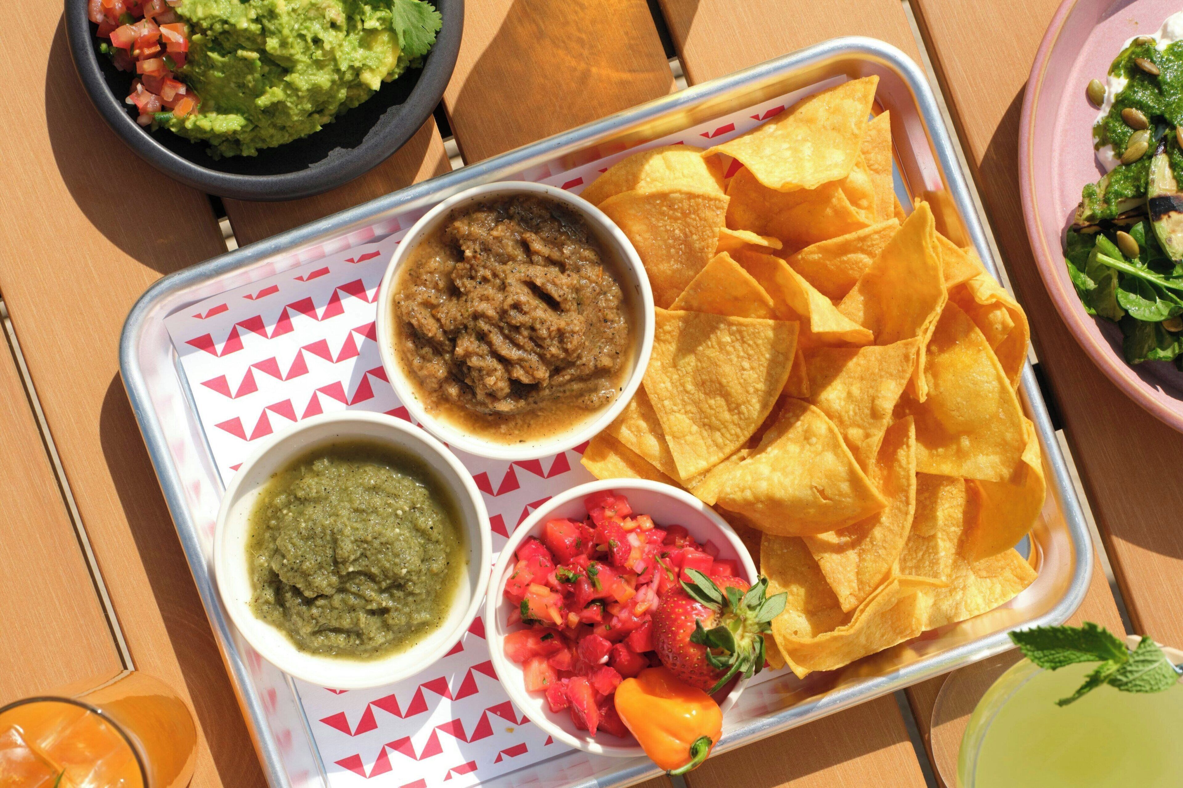 Tarjoilualusta, jossa on nachoja, guacamolea, salsa-kastiketta ja juoma.