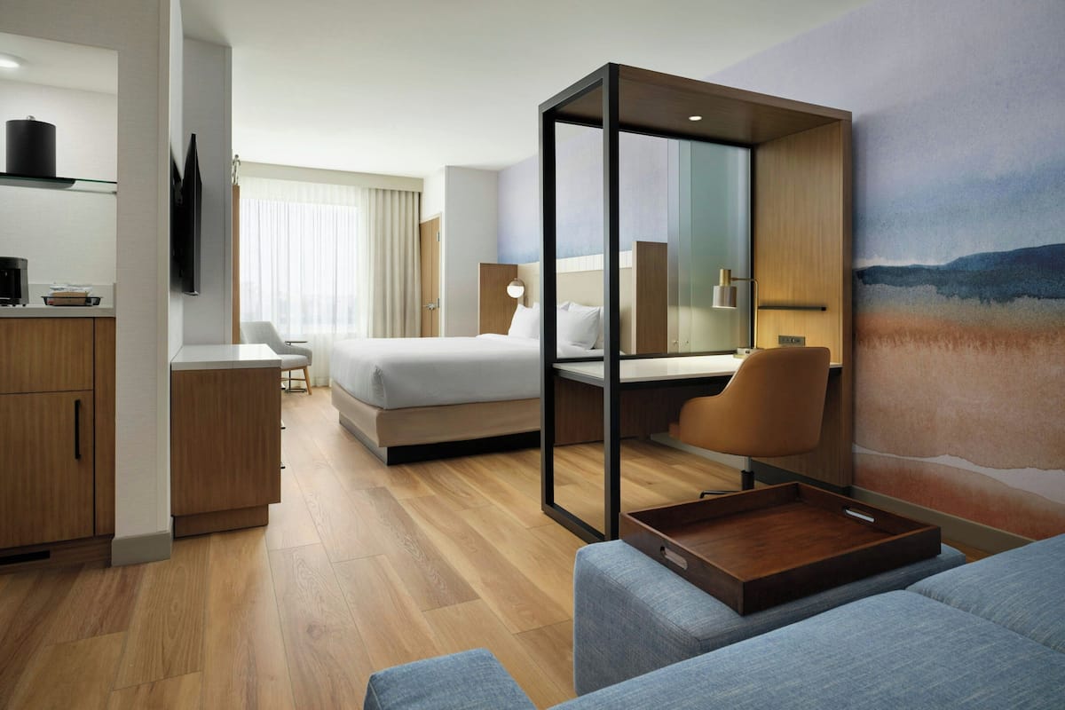 A modern hotel room with a bed, a seating area, a desk, and a TV.