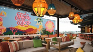 An outdoor seating area with cushioned benches and tables, a mural with the text 'Flora Loca,' and hanging pendant lights. - Springhill Suites By Marriott Ventura Oxnard (Oxnard)
