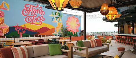 Un espacio al aire libre con bancos acolchados y mesas, un mural con el texto 'Flora Loca', y lámparas colgantes.