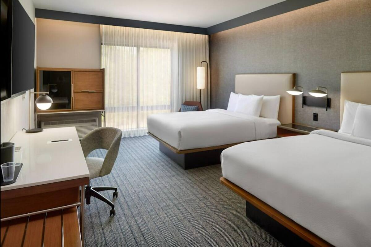 Ein Hotelzimmer mit zwei Betten, einem Schreibtisch, einem Stuhl, einem Fernseher und einem Fenster mit Vorhängen.