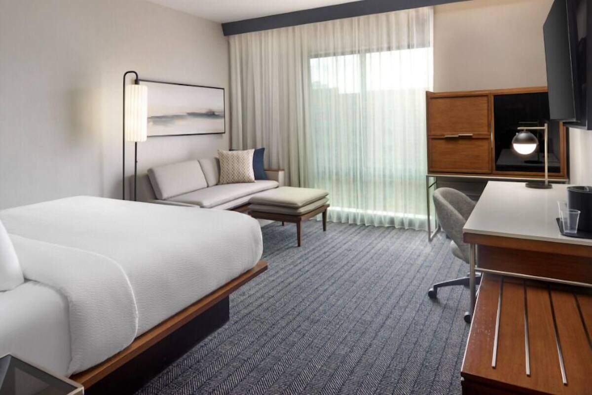 Ein Hotelzimmer mit Bett, Schreibtisch, Stuhl, Fernseher und einem Fenster mit Vorhängen.