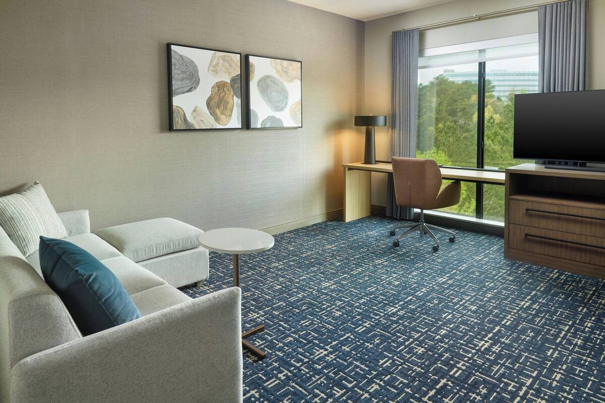 A hotel room with a sofa, a small round table, a TV, and a desk with a chair. There are two framed artworks on the wall and a large window with a view.