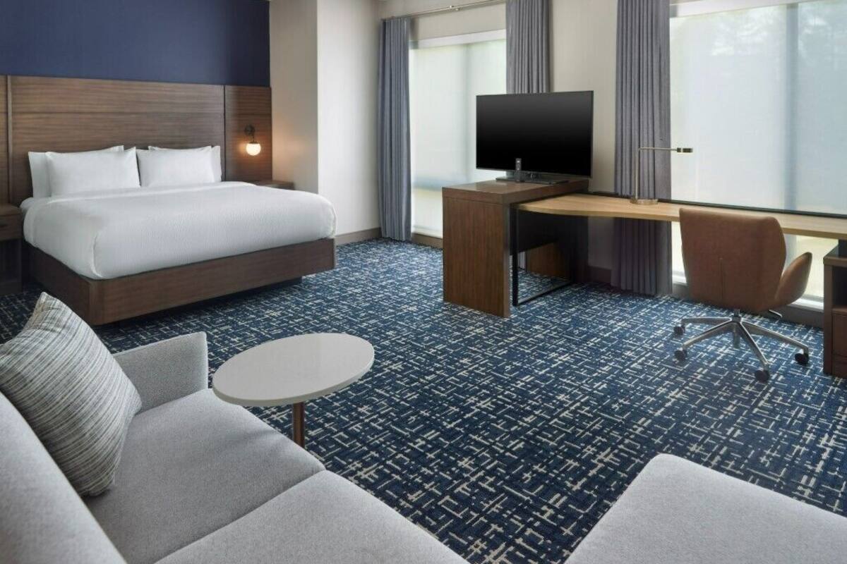 A hotel room with a bed, a desk with a TV, a sofa, and a large window with curtains.