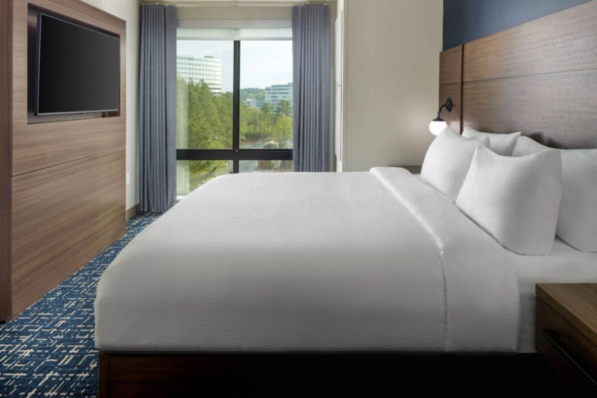 A hotel room with a large bed, a flat-screen TV mounted on the wall, a large window with curtains, and a view of buildings and trees.