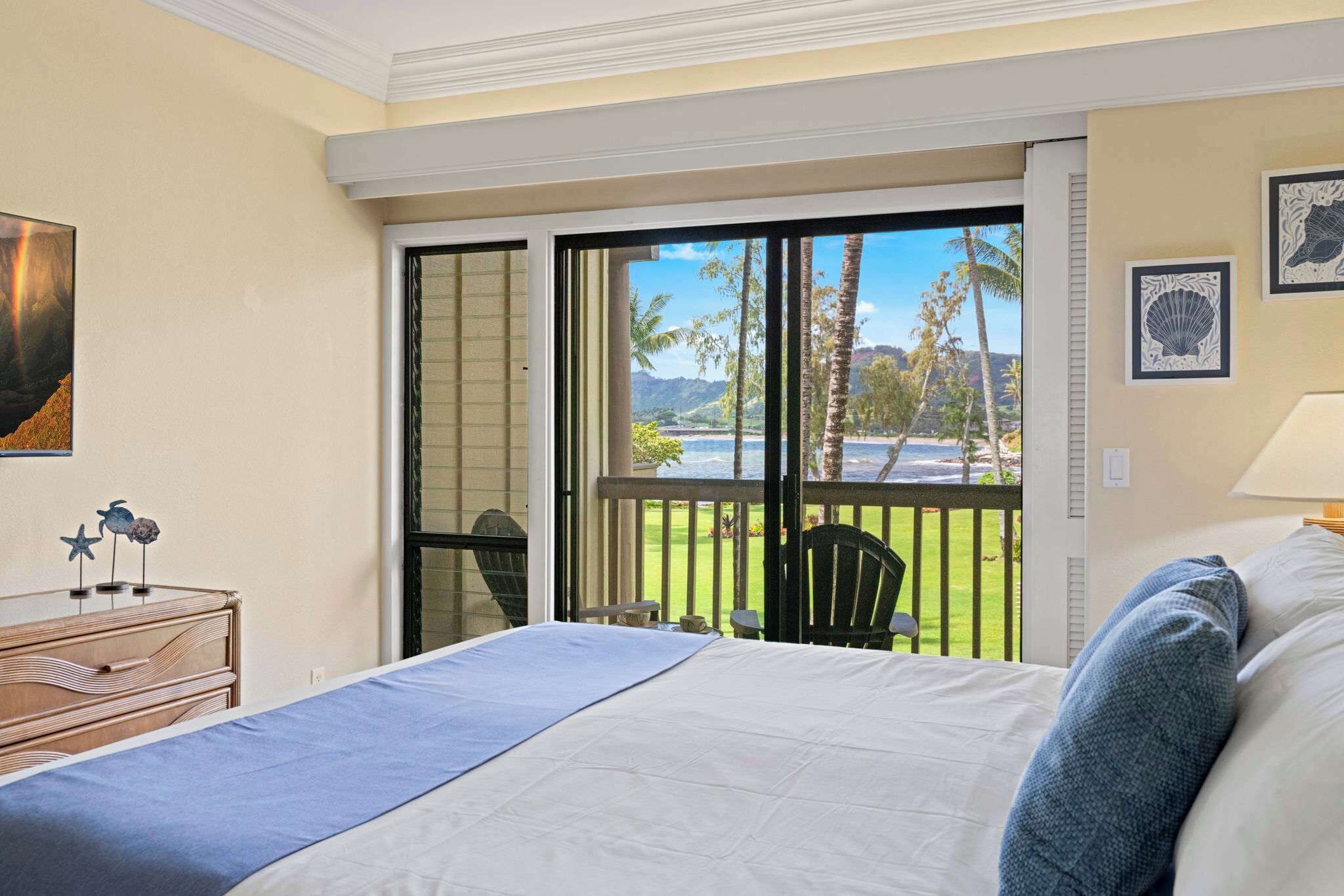 Een slaapkamer met een bed, een dressoir, een stoel en een balkon met uitzicht op de oceaan en palmbomen.
