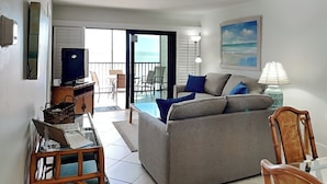 Uma sala de estar com sofá, televisão, abajur e vista para o oceano pelas portas da varanda.