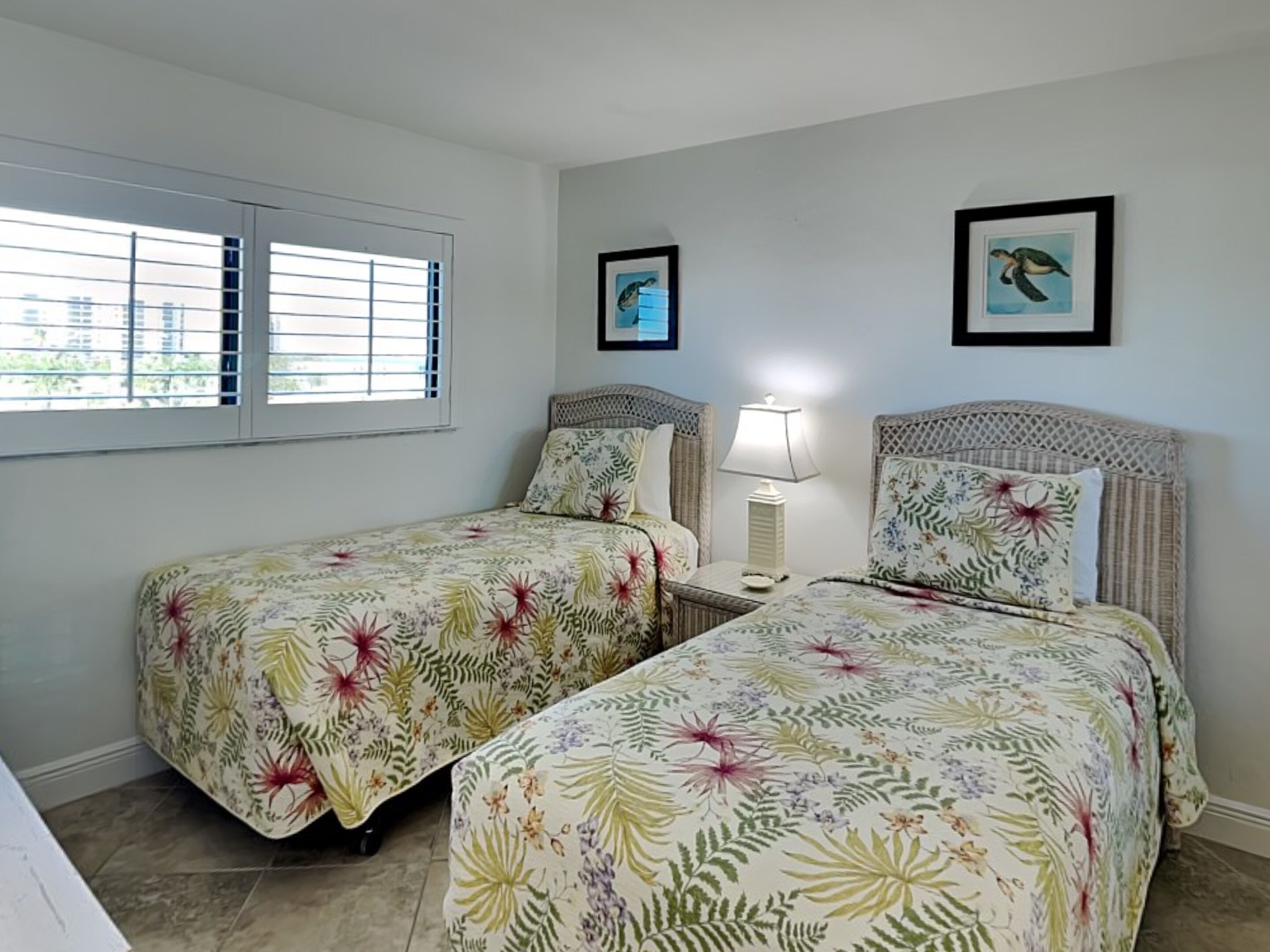 A room with two beds, each with floral bedding, a nightstand with a lamp, and a window with blinds.