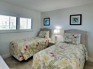 A room with two beds, each with floral bedding, a nightstand with a lamp, and a window with blinds. - Carlos Pointe 422 (Fort Myers Beach)
