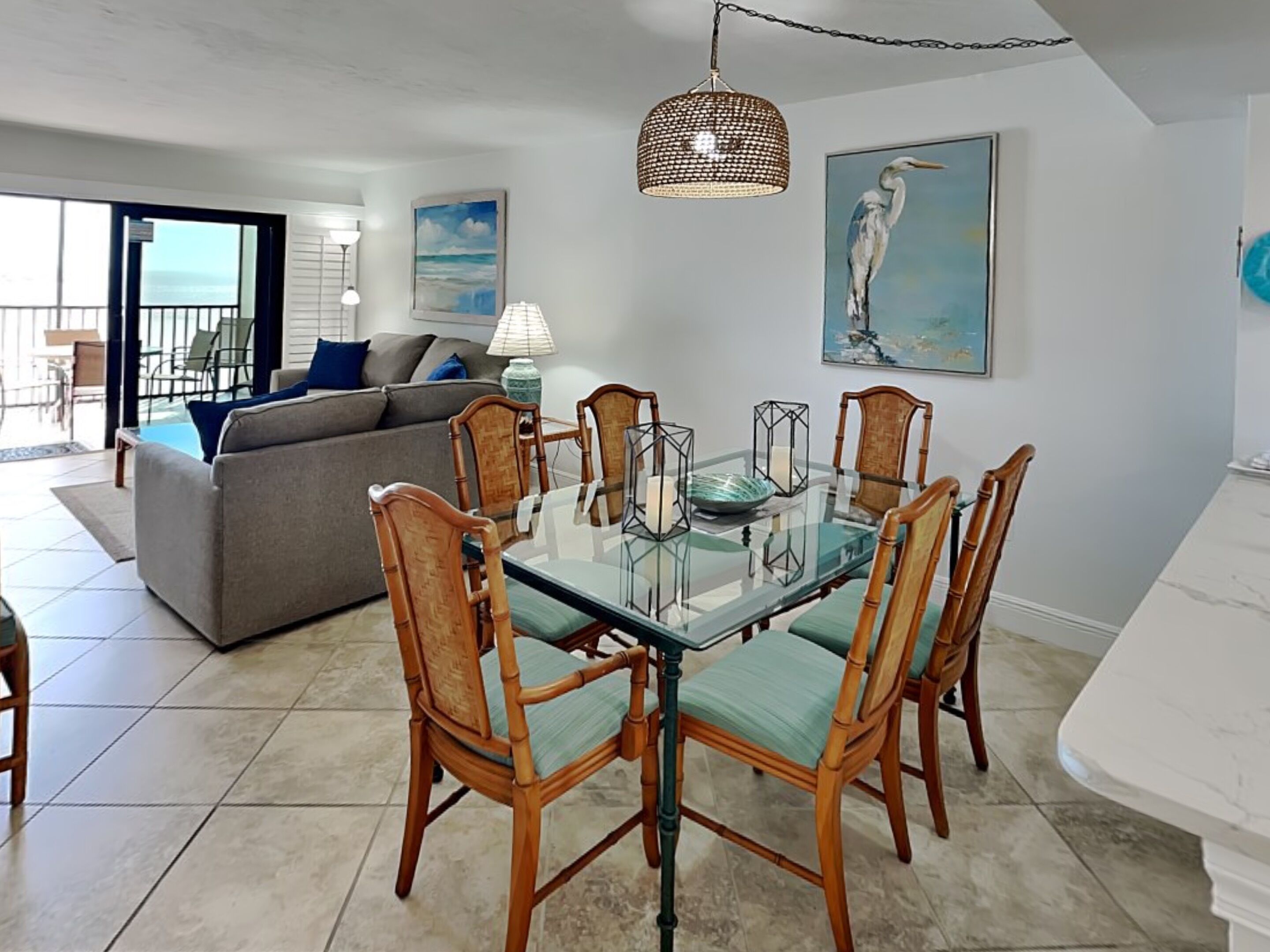 A dining area with a glass table and wooden chairs, a sofa, and a painting of a bird on the wall.