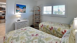 Quarto com duas camas, uma mesinha de cabeceira, cômoda, janela com persianas, quadro na parede e área de cozinha visível por uma porta aberta.