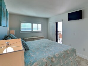 Ein Schlafzimmer mit einem Bett, einem Schminktisch, einem Fenster mit Jalousien, einem an der Wand montierten Fernseher und einem Balkon mit einem Stuhl.