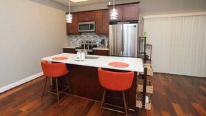 Een moderne keuken met houten kastjes, roestvrijstalen apparatuur en een centraal eiland met twee oranje barstoelen.