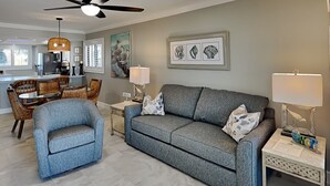 Un salon avec un canapé gris, un fauteuil bleu, un coin repas avec des chaises en osier et un ventilateur de plafond.