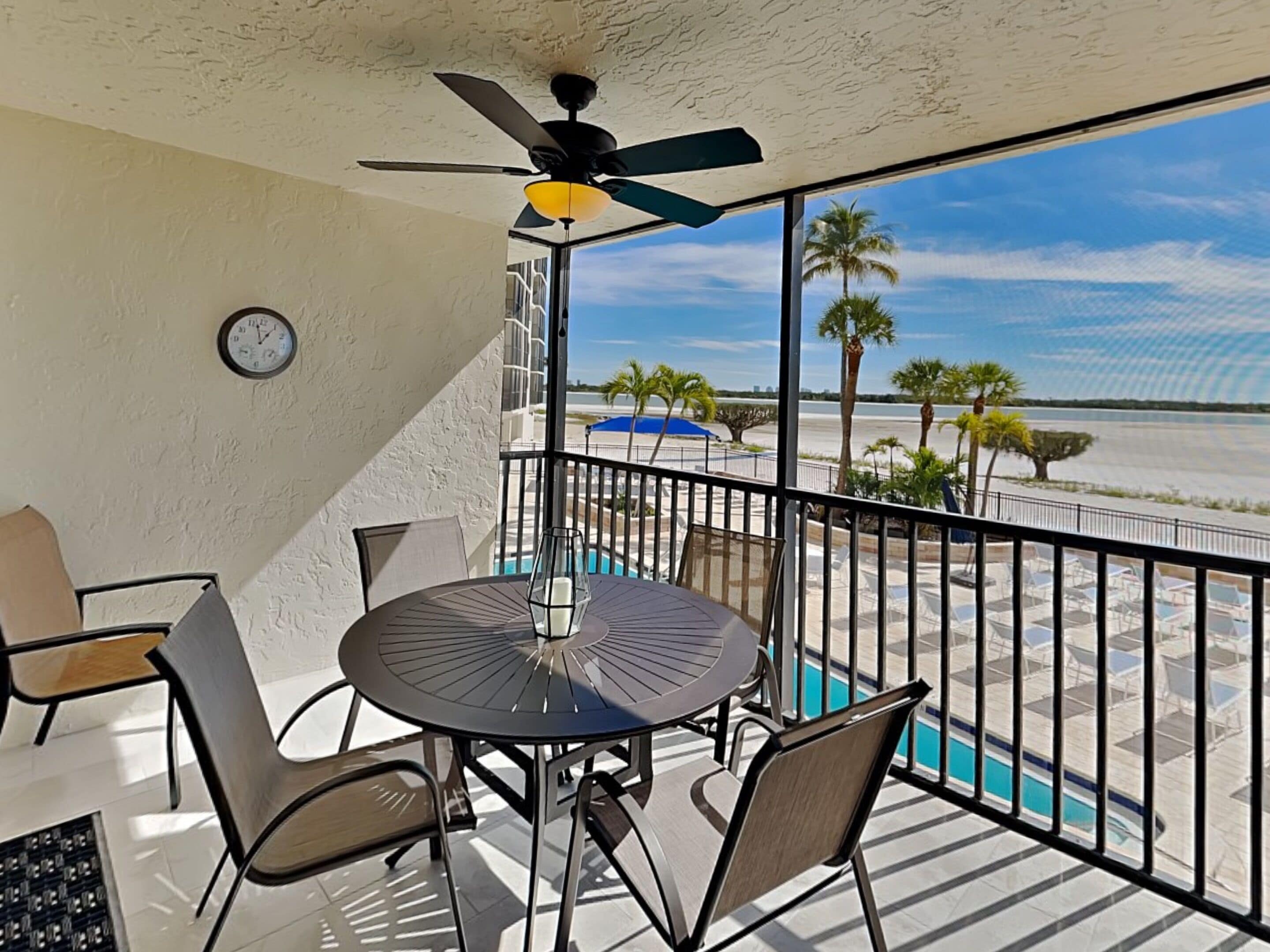 A balcony with a table and chairs, a ceiling fan, and a view of the beach and palm trees.