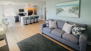Een moderne woonkamer met een grijze bank, een salontafel en een schilderij van een strandlandschap. Er is een keukenhoek met een bar en barkrukken.