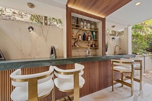 A modern bar area with a marble countertop, three white bar stools with gold accents, and a wooden slatted wall behind the bar. - The Sinatra House- Fully Furnished! (Los Angeles)