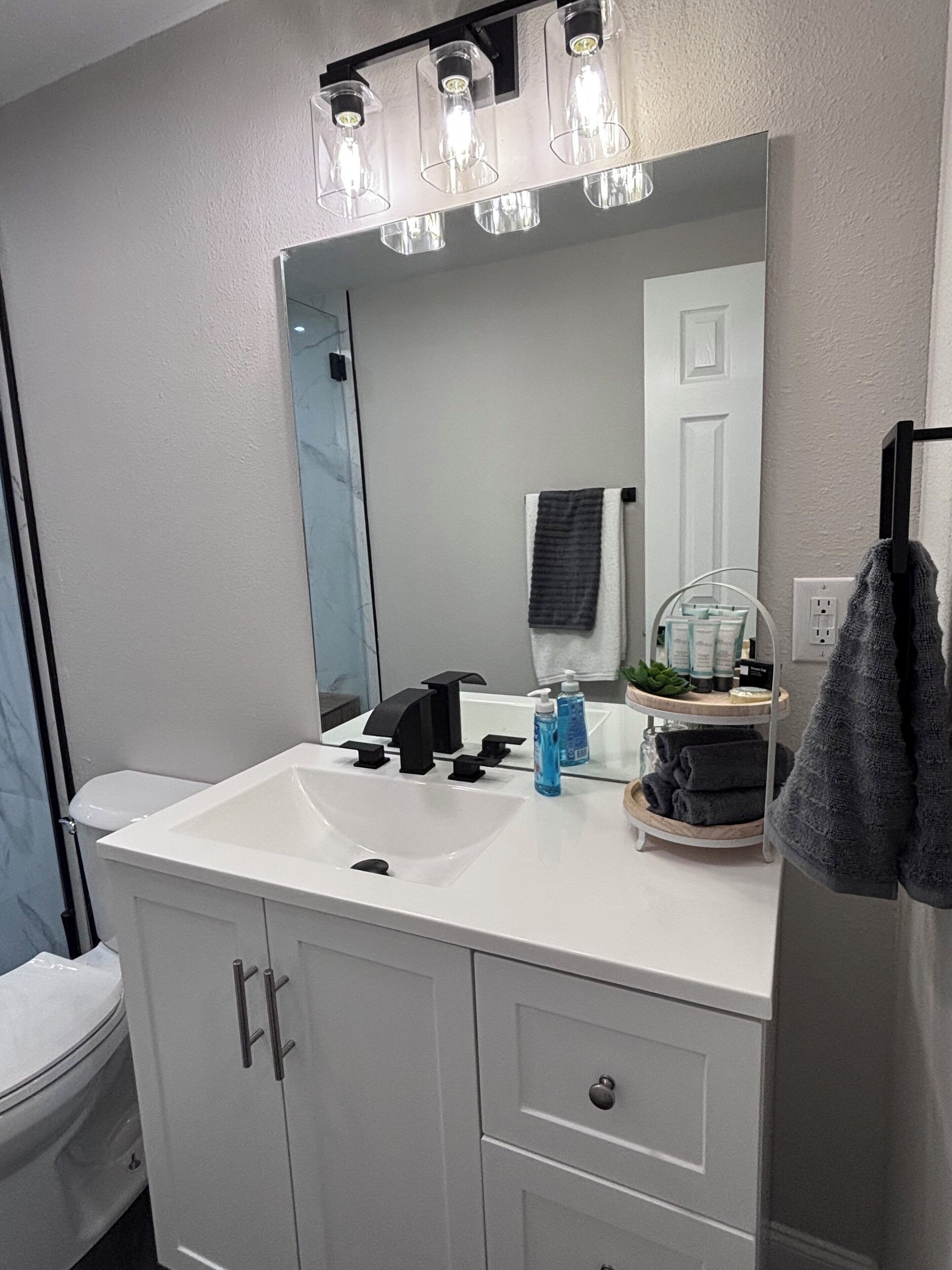 Casa de banho com lavatório branco, torneira preta e espelho em cima. Há produtos de higiene no lavatório e um porta-toalhas na parede.