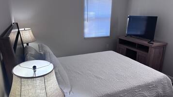 Ein Schlafzimmer mit einem Bett, einer Lampe, einem Fernseher auf einem Holzgestell und einem Fenster mit Jalousien.