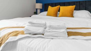 A neatly made bed with white linens and a gold accent, two bright yellow pillows, and a folded white towel on the bed.