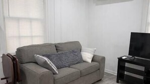 A grey sofa with a patterned pillow, a white pillow, and a black TV stand in a room with white walls and a window with blinds. - Rawson Circle Cottage (Albany)