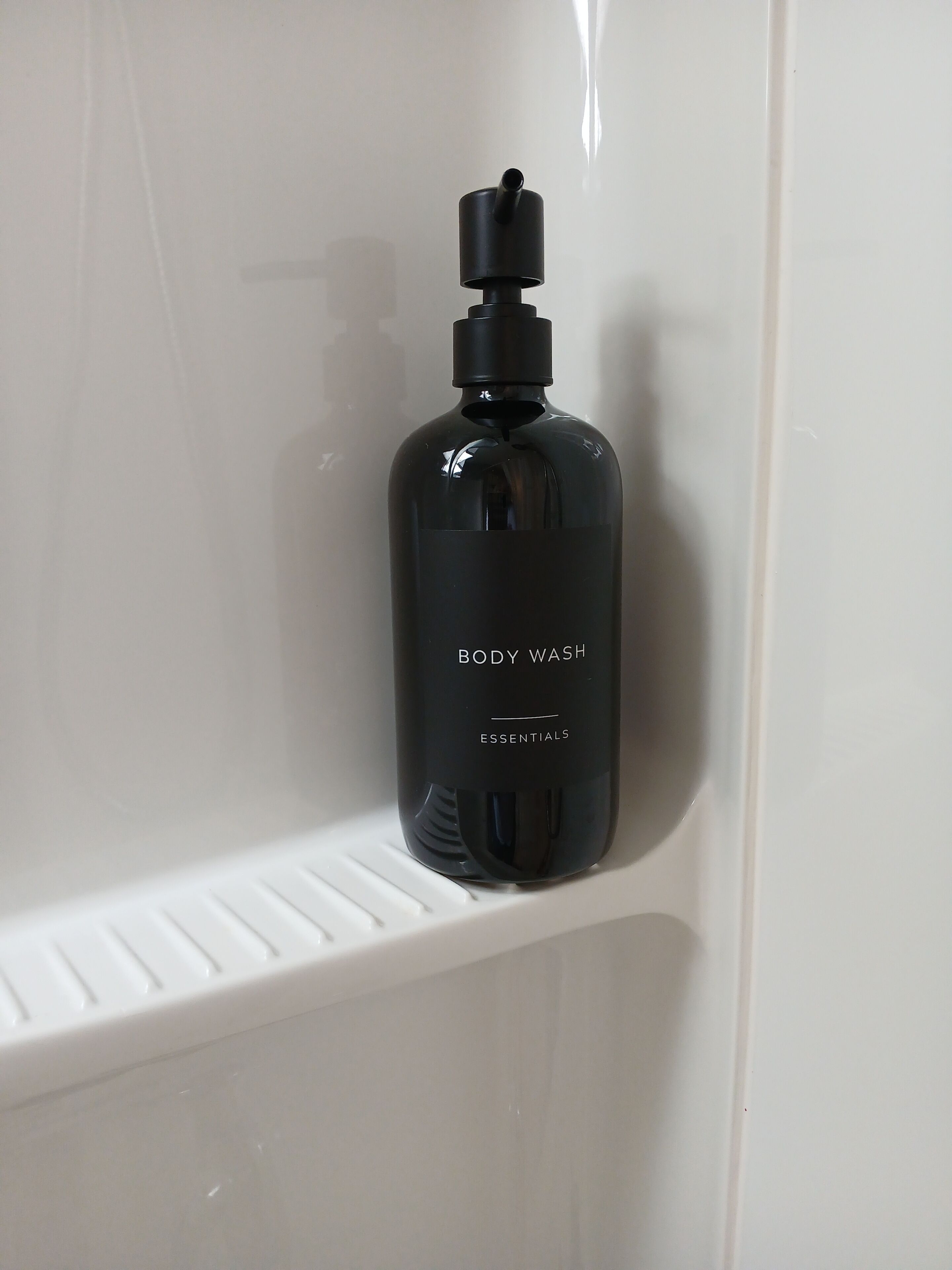 A black bottle of body wash labeled 'BODY WASH ESSENTIALS' on a white shelf.