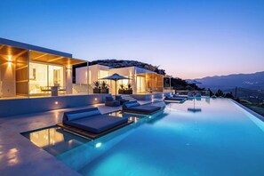 A modern infinity pool with lounge chairs, a well-lit outdoor area, and a view of distant mountains.