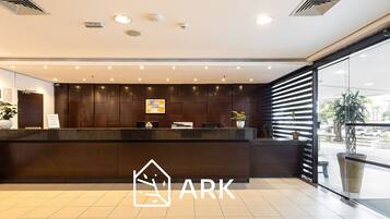 A hotel reception area with a dark reception desk, multiple ceiling lights, and a large window with blinds.