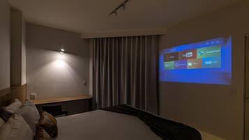 A hotel room with a bed, a wall-mounted projector displaying streaming services, a bedside table, and a curtain.