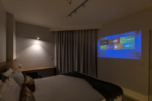 A hotel room with a bed, a wall-mounted projector displaying streaming services, a bedside table, and a curtain. - ARK Houses - Flat Luxo Vista-Mar c/ Piscina na Cobertura (Santos)