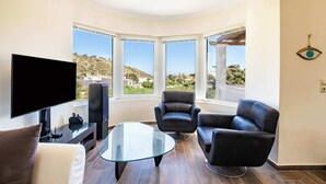Una sala de estar moderna con televisor de pantalla plana, sillones de cuero, una mesa de centro de vidrio y grandes ventanales con vistas al exterior.