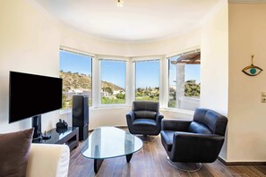 A modern living room with a flat-screen TV, leather chairs, a glass coffee table, and large windows offering a view of the outdoors. - | Zetes Luxury Villa | (Pigadia)